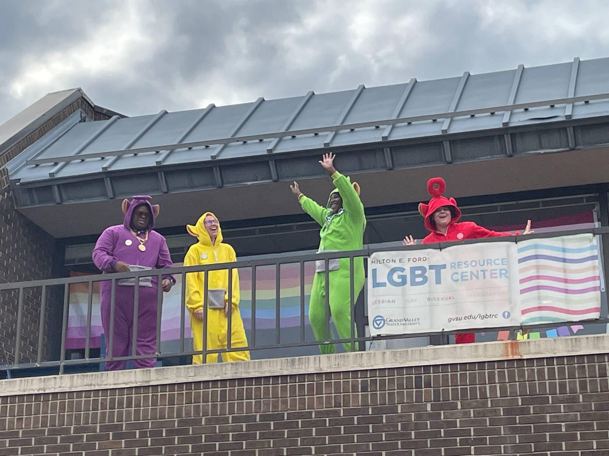 Four people dressed as the Teletubbies in purple, yellow, green, and red costumes pose along an outdoor balcony at GVSU. A cloudy sky and campus building form the background.